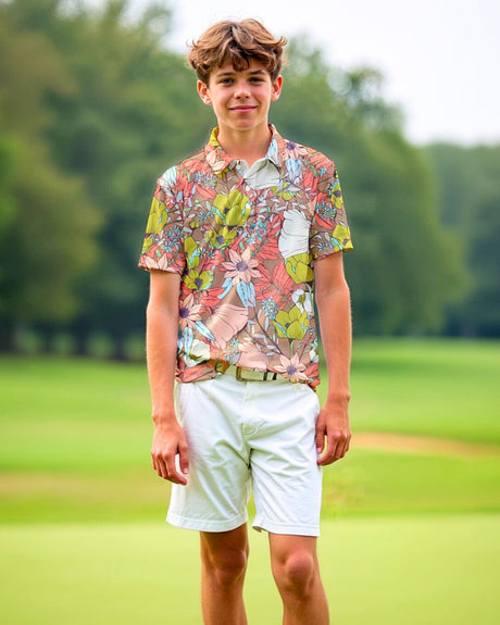 A young man stands on a golf course, smiling and wearing a floral golf polo and white shorts.