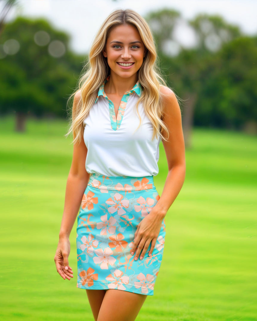 A smiling woman stands on a golf course wearing a white golf polo with a floral-print collar and a matching floral-print skirt.